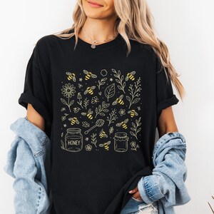 May include: Black t-shirt featuring a golden yellow bee-themed design. The design includes bees, flowers, leaves, honey jars, and a honey dipper. The word "HONEY" is printed on one of the jars. The shirt has short sleeves and a relaxed fit.