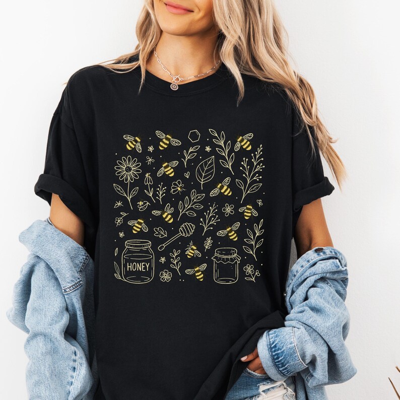 May include: Black t-shirt featuring a golden yellow bee-themed design. The design includes bees, flowers, leaves, honey jars, and a honey dipper. The word "HONEY" is printed on one of the jars. The shirt has short sleeves and a relaxed fit.