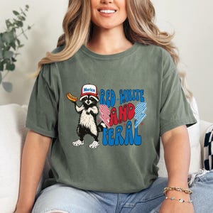 K&ouml;nnte beinhalten: Olivgr&uuml;nes T-Shirt mit einem Waschb&auml;ren, der einen "Merica"-Hut tr&auml;gt und einen Hotdog h&auml;lt. Der Text "Red White and Feral" ist in Rot, Wei&szlig; und Blau gehalten. Der Waschb&auml;r tr&auml;gt blaue und wei&szlig;e Clogs.