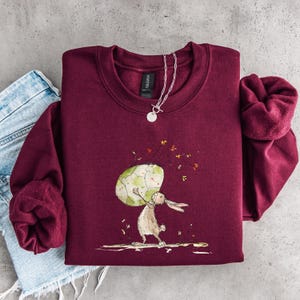 May include: A maroon crewneck sweatshirt featuring a graphic illustration of a light brown rabbit holding a large, patterned green and white egg. The design includes small scattered floral elements and a silver-toned chain necklace resting on the collar.