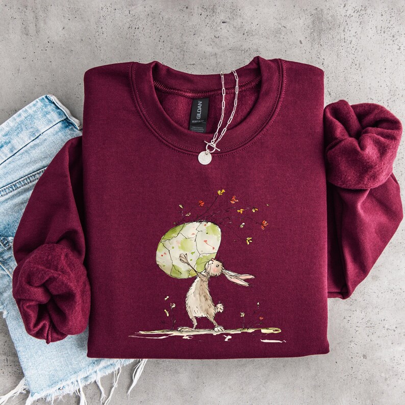 May include: A maroon crewneck sweatshirt featuring a graphic illustration of a light brown rabbit holding a large, patterned green and white egg. The design includes small scattered floral elements and a silver-toned chain necklace resting on the collar.