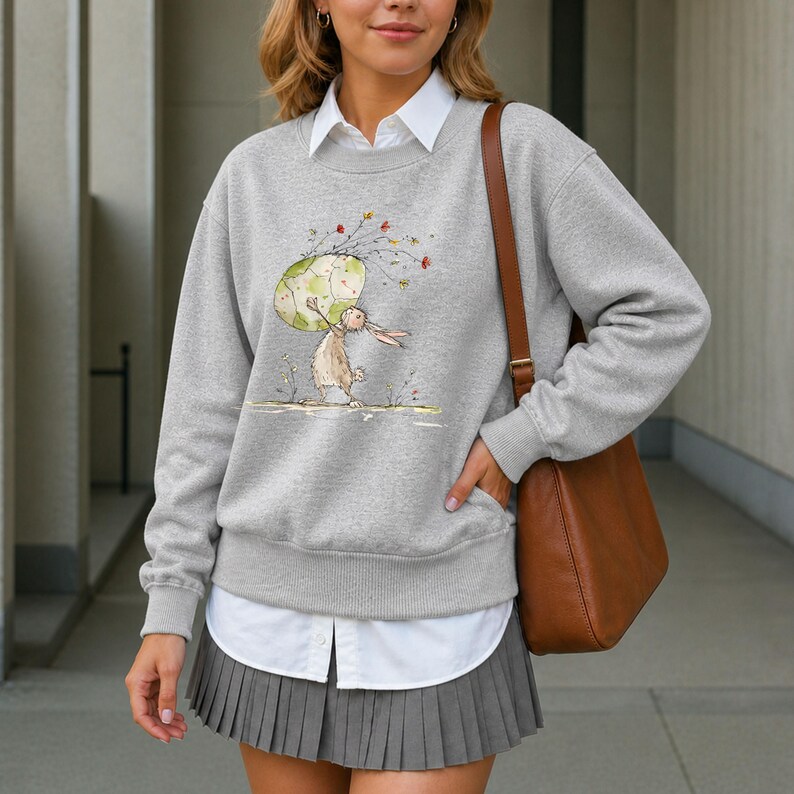 May include: A heather gray crewneck sweatshirt featuring a watercolor illustration of a rabbit holding a large patterned egg with floral accents. The sweatshirt is layered over a white collared shirt and paired with a gray pleated mini skirt.