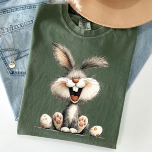 Puede incluir: Camiseta verde oliva con un conejo de dibujos animados sonriendo ampliamente y dos huevos de Pascua decorados. La camiseta está colocada sobre unos vaqueros azules, con un collar dorado y un sombrero beige en el fondo.