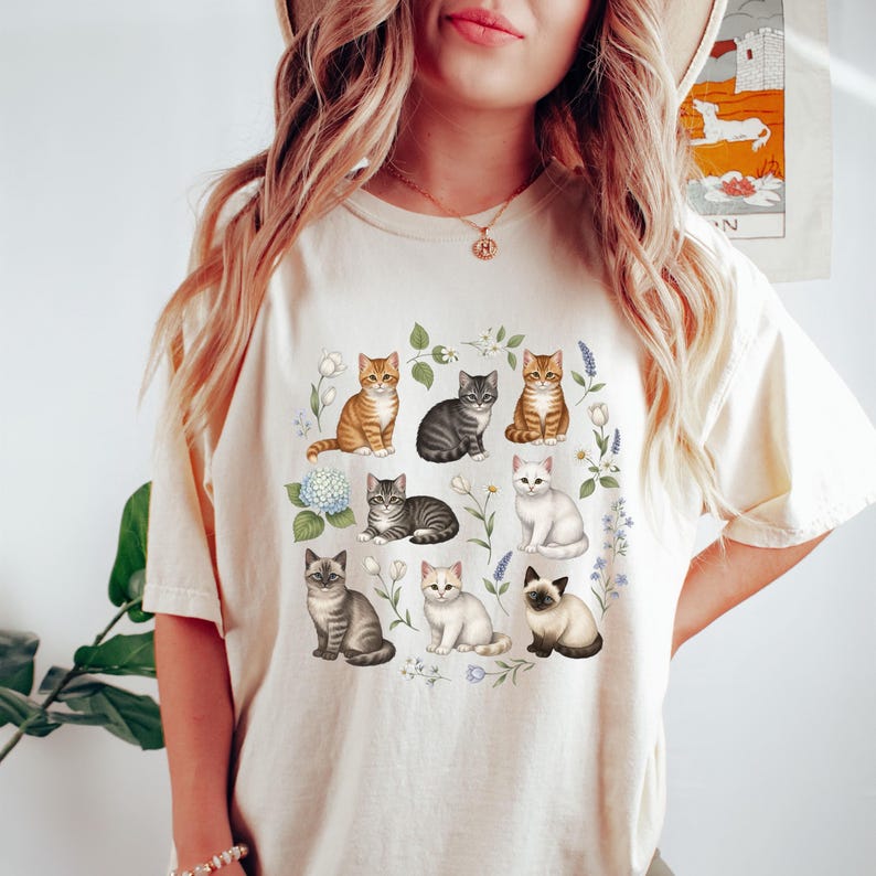 May include: Cream-colored t-shirt featuring a watercolor illustration of nine different cat breeds surrounded by floral elements. The cats are in various poses and colors, including tabby, gray, white, and Siamese. The design is centered on the shirt.