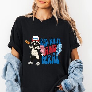 K&ouml;nnte beinhalten: Schwarzes T-Shirt mit einem Waschb&auml;ren, der einen Hut mit der Aufschrift "Merica" tr&auml;gt und einen Hotdog h&auml;lt. Der Text "Red White and Feral" ist in Rot und Blau gehalten, mit Blitz-Grafiken. Der Waschb&auml;r tr&auml;gt wei&szlig;e Clogs.