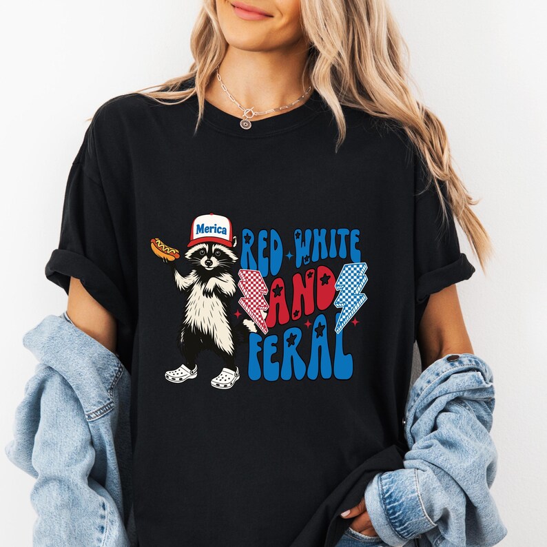 K&ouml;nnte beinhalten: Schwarzes T-Shirt mit einem Waschb&auml;ren, der einen Hut mit der Aufschrift "Merica" tr&auml;gt und einen Hotdog h&auml;lt. Der Text "Red White and Feral" ist in Rot und Blau gehalten, mit Blitz-Grafiken. Der Waschb&auml;r tr&auml;gt wei&szlig;e Clogs.
