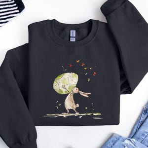 May include: A black crewneck sweatshirt featuring a graphic illustration of a brown rabbit standing on its hind legs while holding a large, pale green, cracked egg above its head, surrounded by small, colorful floral accents and stems.