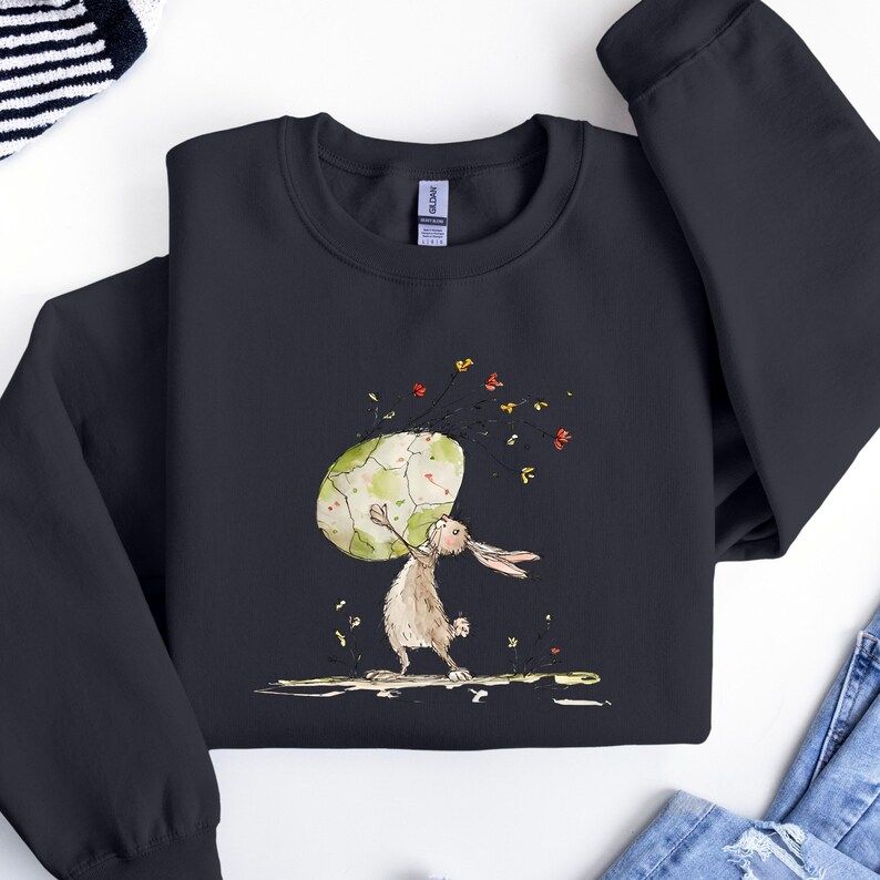 May include: A black crewneck sweatshirt featuring a graphic illustration of a brown rabbit standing on its hind legs while holding a large, pale green, cracked egg above its head, surrounded by small, colorful floral accents and stems.