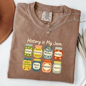 May include: A brown t-shirt with the phrase "History is My Jam" and illustrations of jars labeled with historical periods. The shirt also has a Comfort Colors tag and a silver necklace.