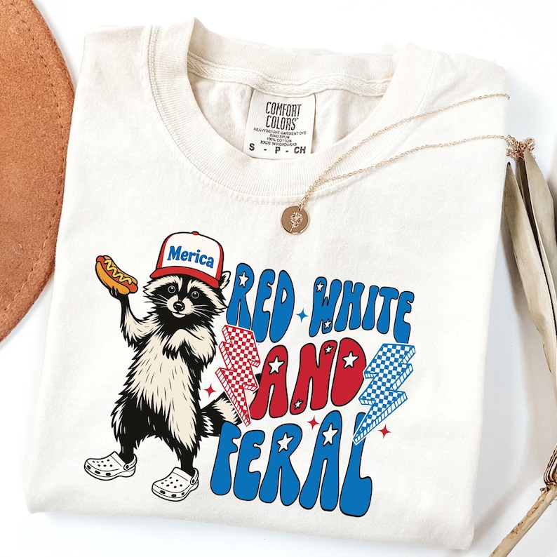 May include: Cream-colored t-shirt featuring a raccoon wearing a hat that says "Merica" and holding a hot dog. The shirt has the text "Red White and Feral" in red and blue, with lightning bolt graphics. A gold necklace is draped on the shirt.