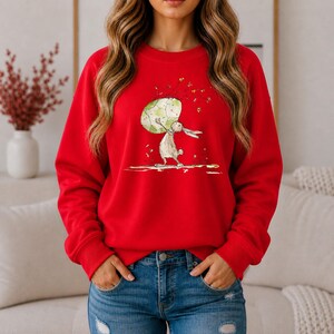 May include: A bright red long-sleeve sweatshirt featuring a central graphic illustration of a light-colored rabbit standing on its hind legs while holding a large, textured green and white patterned sphere above its head with small gold accents.