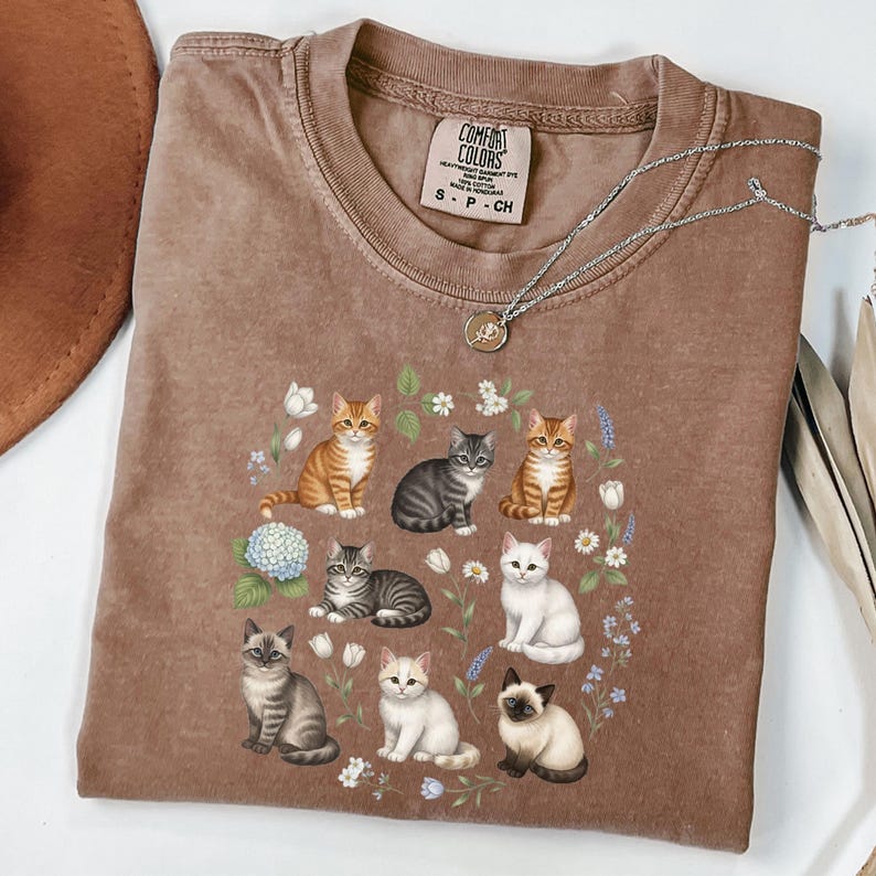 May include: Brown Comfort Colors t-shirt featuring a floral wreath design with various cat illustrations. The cats are in different poses and colors, surrounded by flowers and foliage. A silver necklace with a pendant is on top of the shirt.