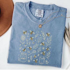 May include: A blue Comfort Colors t-shirt with a bee-themed design. The graphic includes bees, flowers, leaves, and honey jars. A gold necklace with a pendant rests on the shirt. The shirt size is small.