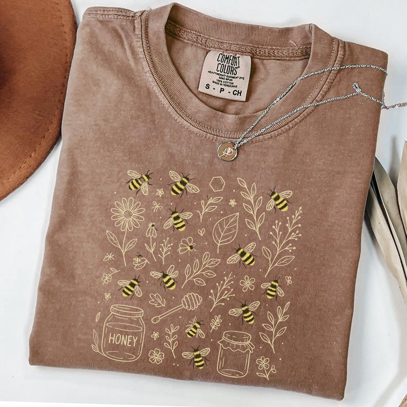 May include: A brown Comfort Colors t-shirt with a bee and honey-themed graphic. The design includes bees, flowers, leaves, and honey jars. A silver necklace with a small pendant is draped across the shirt. The shirt is size S-P-CH.