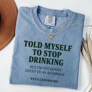 Könnte beinhalten: Ein hellblaues Comfort Colors T-Shirt mit dem Text "TOLD MYSELF TO STOP DRINKING" in Dunkelgrün. Das Shirt hat auch den Text "BUT I'M NOT GONNA LISTEN TO AN ALCOHOLIC" und den Hashtag "#STILLDRINKING". Eine goldene Halskette liegt auf dem Shirt.