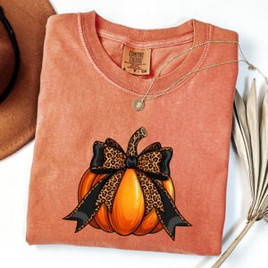 Può includere: T-shirt arancione Comfort Colors con grafica di zucca, fiocco nero e dettagli animalier. Una collana d'oro con un ciondolo è drappeggiata sulla maglietta. La maglietta ha un'etichetta con la scritta "Comfort Colors".