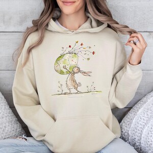 May include: A beige hooded sweatshirt featuring a central graphic illustration of a brown rabbit holding a large, light green patterned egg. The design includes delicate, thin-lined floral stems and small colorful flowers extending upward from the egg.