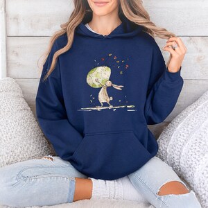 May include: A navy blue hooded sweatshirt featuring a graphic illustration of a brown rabbit holding a large, speckled, light green egg. The design includes small, colorful floating leaves and petals surrounding the rabbit and egg.
