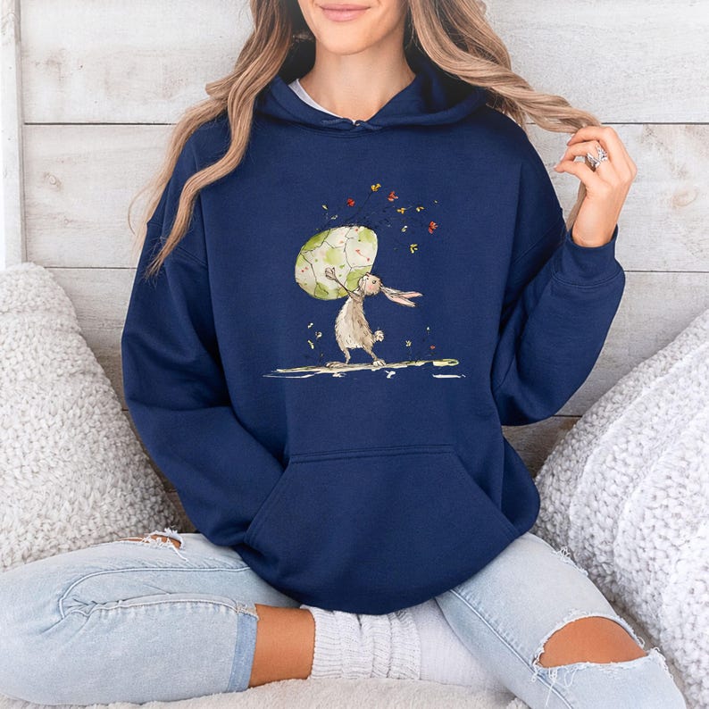 May include: A navy blue hooded sweatshirt featuring a graphic illustration of a brown rabbit holding a large, speckled, light green egg. The design includes small, colorful floating leaves and petals surrounding the rabbit and egg.