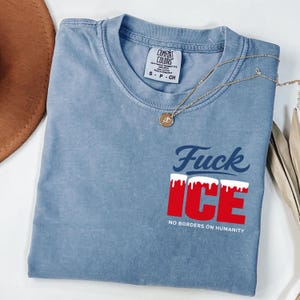 May include: A blue Comfort Colors t-shirt with the text "Fuck ICE" in navy blue and red, with white accents. The shirt also has the text "NO BORDERS ON HUMANITY". A gold necklace with a pendant is draped across the shirt.