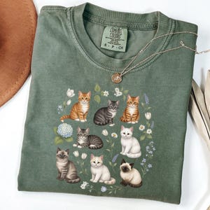 May include: A sage green Comfort Colors t-shirt featuring a graphic of various cat breeds surrounded by flowers. The cats are in different poses and colors, including orange, gray, and white. A gold necklace and a brown hat are also visible.
