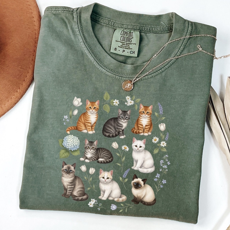 May include: A sage green Comfort Colors t-shirt featuring a graphic of various cat breeds surrounded by flowers. The cats are in different poses and colors, including orange, gray, and white. A gold necklace and a brown hat are also visible.