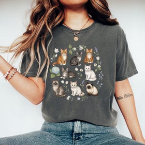 May include: Dark grey t-shirt featuring a floral arrangement of various cat breeds. The cats are in different colors and patterns, surrounded by white flowers, lavender, and a blue hydrangea. The shirt is a comfortable, casual style.