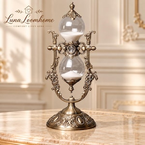 May include: An antique-style brass hourglass featuring a rotating frame with intricate floral scrollwork and a central clear crystal accent. The glass bulbs contain white sand, and the base is decorated with embossed floral patterns.