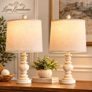 May include: Two table lamps with off-white, textured fabric drum shades and distressed white, turned-wood style bases. The lamps feature a classic silhouette and emit a warm glow. The brand name Luna Loomhome is visible in the upper left corner.