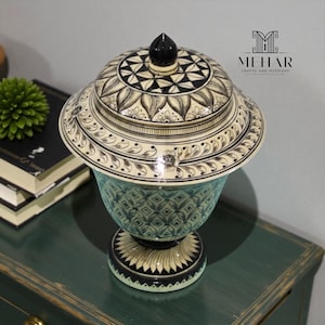 Puede incluir: Un jarrón decorativo de cerámica con tapa. El jarrón tiene un cuerpo azul claro con un patrón geométrico, un borde y tapa color crema con diseños florales negros y un remate negro. El jarrón está sobre una superficie verde oscuro.