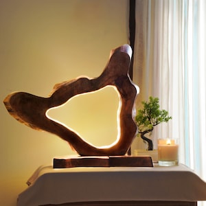Puede incluir: Una escultura de madera única con una gran abertura irregular iluminada por una luz suave y cálida. La escultura se asienta sobre una base de madera rectangular. Un pequeño bonsái y una vela encendida están en el fondo.