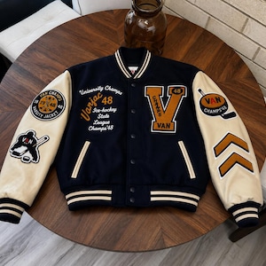 May include: A navy blue varsity jacket with cream-colored sleeves, featuring embroidered patches and text including "University Champs 48", "Vanjac 48", "Ice-hockey State League Champs 48", and a large "V" on the chest with "VAN" and "48" details.