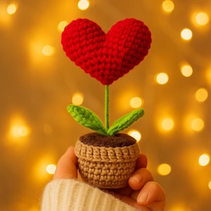 以下が含まれることがあります： 赤いハート型の花、緑の葉、茶色の茎を持つ手作りのかぎ針編みの花。小さな茶色のかぎ針編みの鉢に植えられています。背景は、暖かくぼやけた金色の光のボケです。