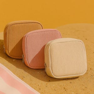 May include: Three corduroy cosmetic bags in shades of tan, pink, and cream, with vertical ribbed texture and a zip closure. The bags are arranged on a sandy surface with a striped towel in the foreground.