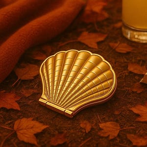 Puede incluir: Un espejo compacto dorado en forma de concha marina descansa sobre un lecho de hojas otoñales. Un vaso de limonada con una rodaja de limón y un libro abierto están cerca. Una manta de color óxido está en el fondo. La escena evoca un ambiente otoñal.