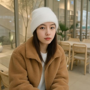 May include: A white knit beanie hat. The hat is worn with a brown fleece jacket and a white shirt. The background is a blurred indoor setting with tables and chairs.