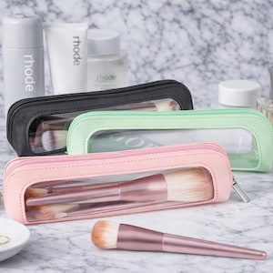 May include: Three makeup brush cases in black, mint green, and pink, each with a clear window, containing makeup brushes. Other beauty products, including a perfume bottle, are also visible.