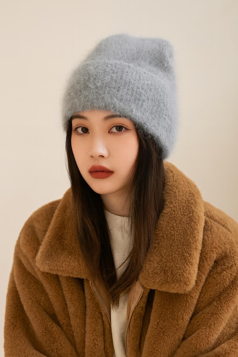 May include: A light blue, fuzzy beanie hat. The hat is worn by a person wearing a brown, fuzzy coat. The coat has a collar and a zipper. The person is looking at the camera.