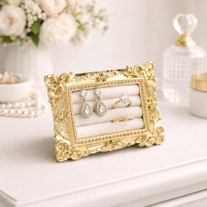 May include: A gold-colored, ornate picture frame displaying jewelry. The frame holds a pair of teardrop earrings, a diamond ring, and a thin band ring. The frame sits on a white surface, with a vase of flowers in the background.