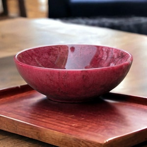 Könnte beinhalten: Eine tiefrote Schüssel steht auf einem passenden Holztablett. Die Schüssel hat eine glatte, glänzende Oberfläche und das Tablett hat einen satten, rötlich-braunen Farbton. Die Gegenstände stehen auf einer Holzoberfläche und erzeugen eine warme, einladende Ästhetik.