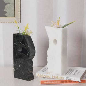 May include: Two marble vases, one black and one white, each with a unique, sculpted design. The black vase has a dark green and white marbled pattern. The white vase has a circular cut-out. Both vases hold small floral arrangements.
