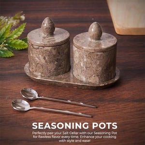 Pode incluir: Um conjunto de dois potes de temperos com tampas combinando, com um padrão de mármore, repousando em uma bandeja oval. Duas pequenas colheres de prata são colocadas na frente. O texto "SEASONING POTS" é exibido abaixo.