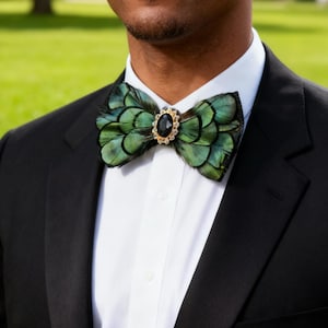 Puede incluir: Una pajarita de plumas verdes con un centro de gema negra y detalles dorados. La pajarita se lleva con una camisa blanca y una chaqueta de traje negra. El fondo es verde borroso.