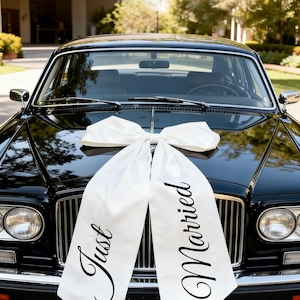 May include: A black vintage car with a large white bow on the hood. White ribbons with the words "Just Married" are draped over the front of the car. The car is parked outside a building.