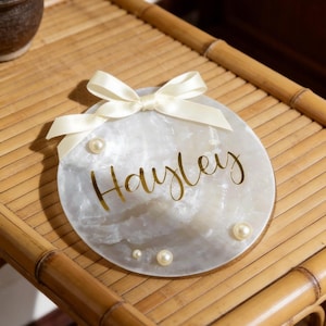 May include: A circular white marbled plaque featuring the name Hayley in gold script lettering. The plaque is decorated with a cream-coloured satin ribbon bow at the top and four small white pearls of varying sizes attached to the surface.
