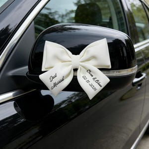 May include: A white bow tied to a black car wing mirror. The bow is inscribed with "Just Married" and the names "Owen & Eloan" with the date "02-11-2026". The car is black, with a blurred background of trees and sky.