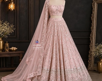 May include: A pale pink lehenga set featuring a sleeveless crop top and a floor-length skirt, both heavily embellished with silver sequins and mirror work. A matching sheer dupatta with a decorative border is draped over the shoulder.