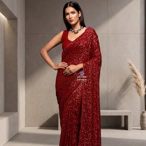 May include: A woman in a red sequined saree. The sleeveless top and draped fabric are covered in shimmering sequins. The saree is a deep red colour, and the woman is wearing a necklace and ring. The image is taken indoors.