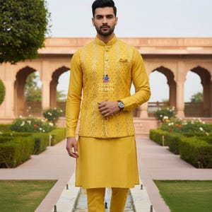 Op de afbeelding: Een mosterdgeel driedelig ensemble. De outfit bestaat uit een shirt met lange mouwen, een vest met ingewikkelde borduursels en een bijpassende broek. De outfit wordt gecompleteerd met goudkleurige schoenen. Er is ook een horloge te zien.