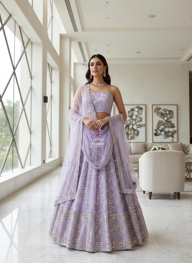 K&ouml;nnte beinhalten: Ein lavendelfarbener Lehenga mit silbernen Verzierungen. Das Set beinhaltet ein Crop-Top, einen Rock und einen transparenten Dupatta. Das Outfit ist mit aufwendigen Details und dem Text "CLOTHS STUDIOS" versehen.